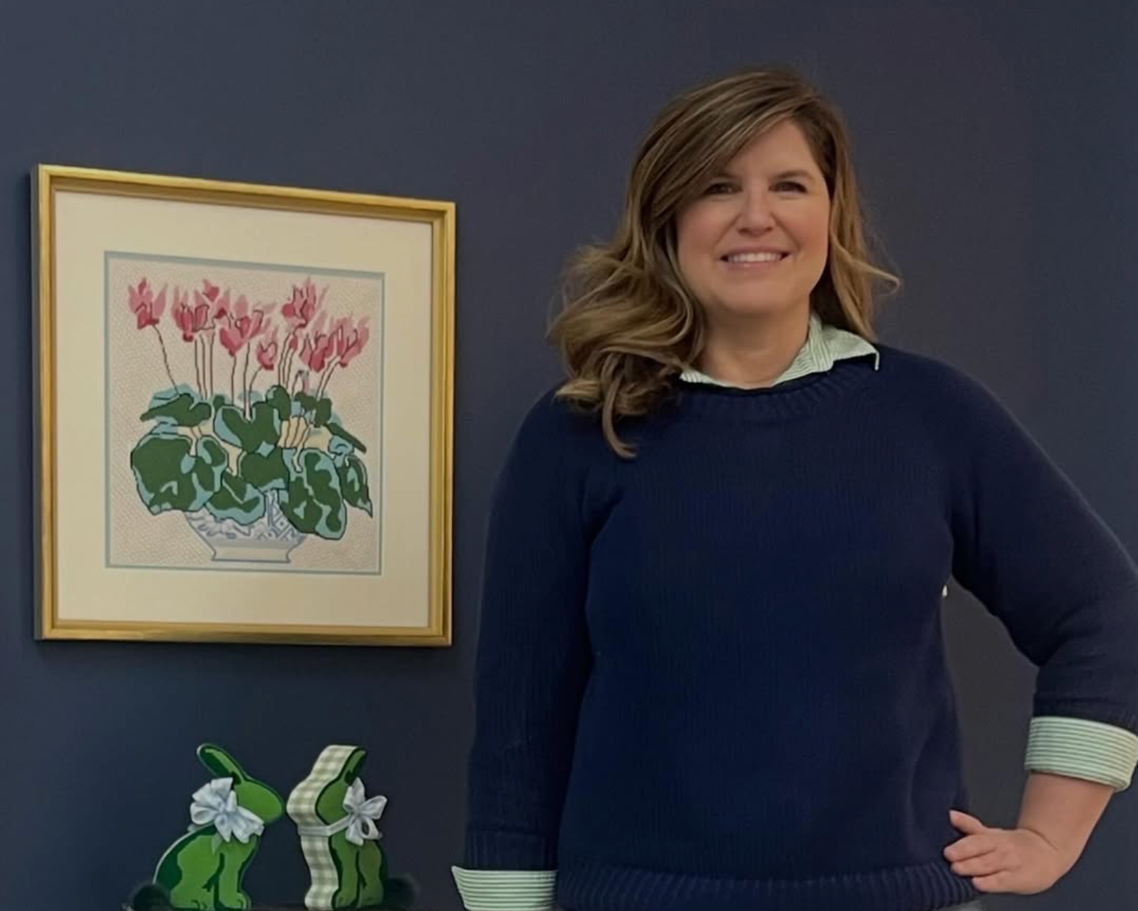 Woman wearing a navy sweater with green cuffs standing in a room with a framed picture of flowers on the wall.